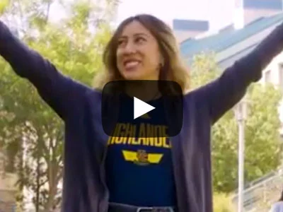 A female student has her arms open in a Y - there is a video play button that is overlaid on top of her. The student is wearing a dark cardigan with a Highlander shirt underneath.