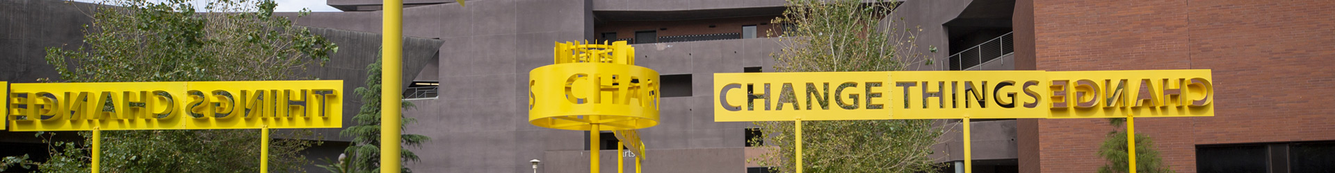 A bright yellow outdoor art installation near the Arts building on the UC Riverside campus featuring the words "CHANGE THINGS" in large, cutout letters against a backdrop of modern university buildings. The text is displayed both forward-facing and mirrored on elevated metal banners and a circular structure.