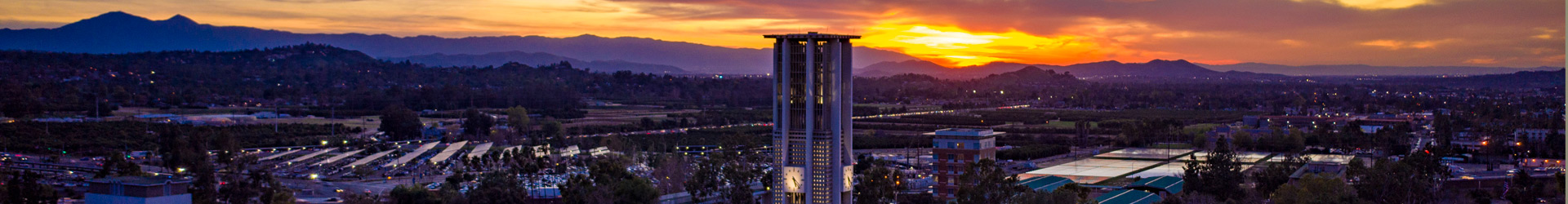 A panoramic sunset view of the University of California, Riverside campus, featuring the illuminated Bell Tower in the center foreground. The background shows city lights and silhouettes of the Box Springs Mountains under a vibrant orange and yellow sky.