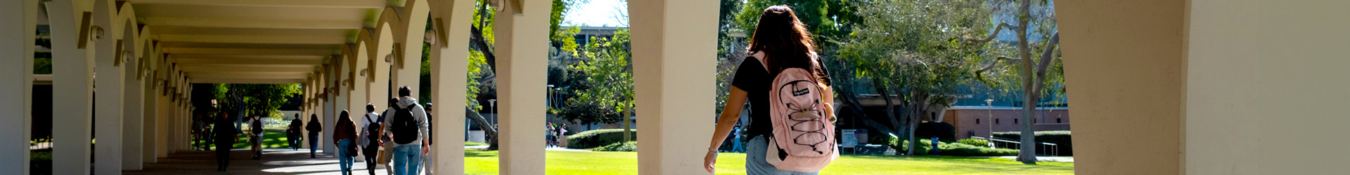 Students walk through the sunlit arches of the Tomas Rivera Library at UC Riverside. In the foreground, a student with a pink backpack faces toward the green lawn.