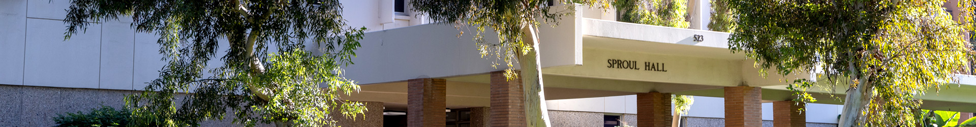 An exterior view of Sproul Hall on the UC Riverside campus. The building features cream-colored architecture and brick pillars partially framed by green leafy trees.