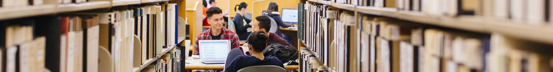 Several students sit at desks with laptops and books in the Orbach Library at UC Riverside. They are framed by two rows of tall bookshelves in the foreground. The atmosphere is academic and focused, highlighting a collaborative study environment.