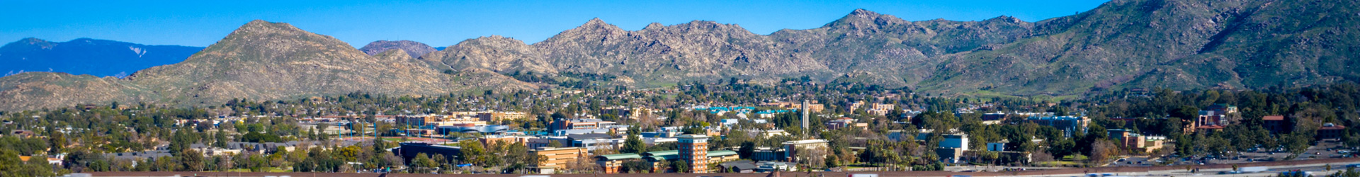A wide panoramic view of a the UC Riverside campus nestled at the base of rugged, rocky mountains under a clear blue sky. The landscape features a mix of academic buildings, green spaces, and palm trees stretching across the valley floor.