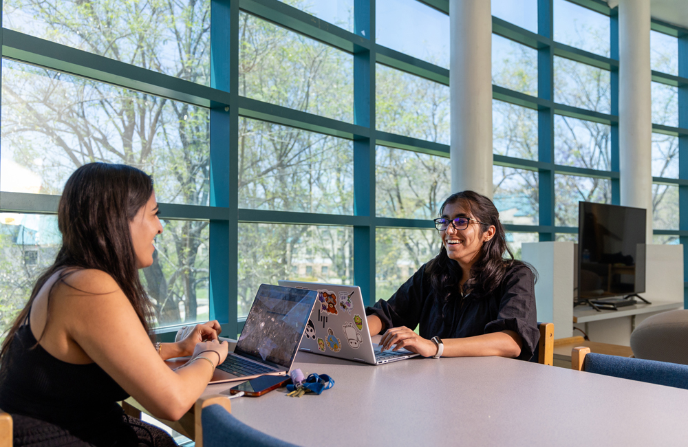 Two students sit across from each other at a table in a bright, modern library or common area, smiling and talking while working on their laptops. Large windows in the background look out onto a green campus with leafy trees.