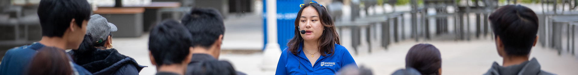 A UCR student guide with a headset microphone speaks to a tour group outdoors on campus.