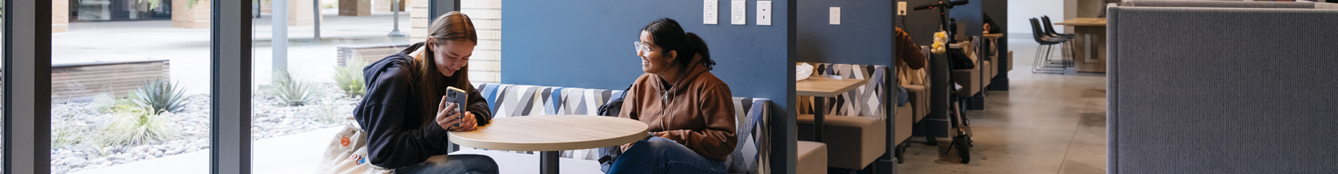 Two female students are sitting together in a modern and brightly lit student lounge. One student smiles as she shares something on her smartphone with her friend, who looks on with a happy, engaged expression. This scene captures a candid moment of friendship and relaxation on campus.