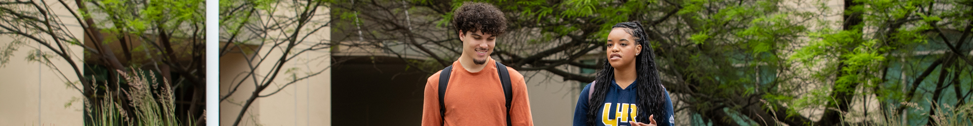 Two students, a young man and woman, walk together on a tree-lined campus path on the UCR campus. The woman, in a UCR sweatshirt, is actively in conversation while the man listens with a friendly smile. This image captures a moment of student connection and daily life on campus.