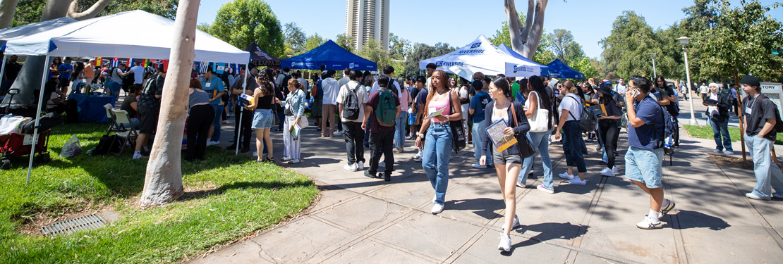 A diverse group of students explores a vibrant UC Riverside transfer student event on campus. Under sunny skies, attendees walk along a paved walkway lined with informational tents to connect with university representatives.
