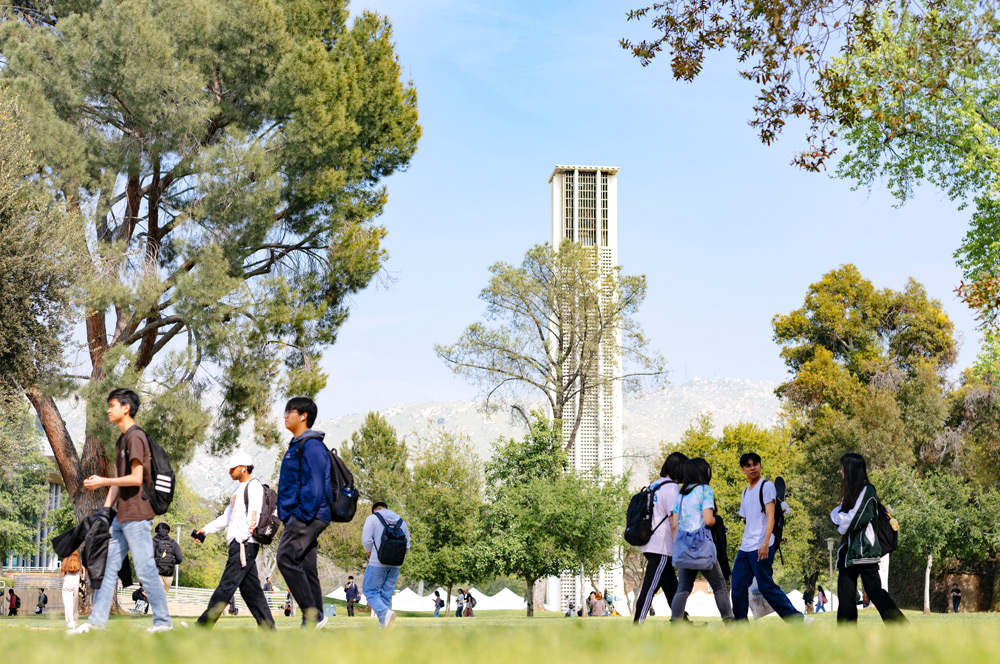 Students walk across a sprawling green lawn on the scenic UC Riverside campus under a clear blue sky. In the background, the iconic UCR Bell Tower rises above the trees, representing a central landmark of the university.