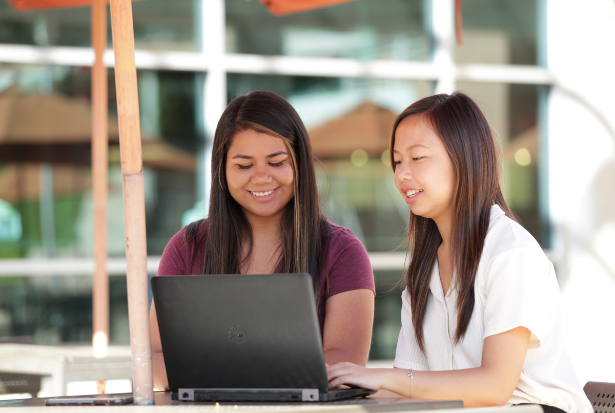 Two smiling female students work together on a laptop at a sunny outdoor table on the UCR campus.