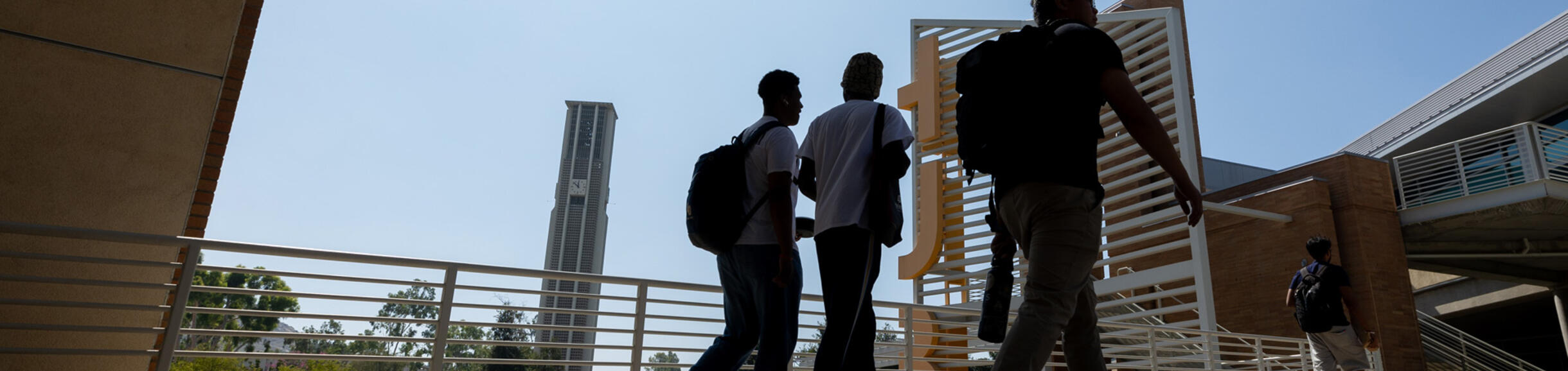 Silhouetted UCR students walk along a path toward the Highlander Union Building (HUB), with the Bell Tower in the background.