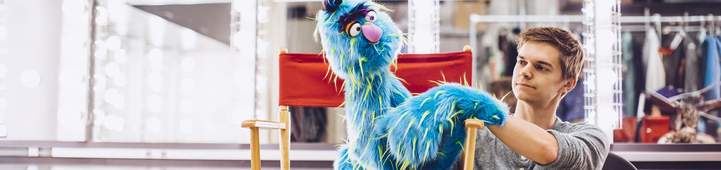 A man adjusts a shaggy blue monster puppet. The background features mirrors and clothing racks, suggesting a theater setting.