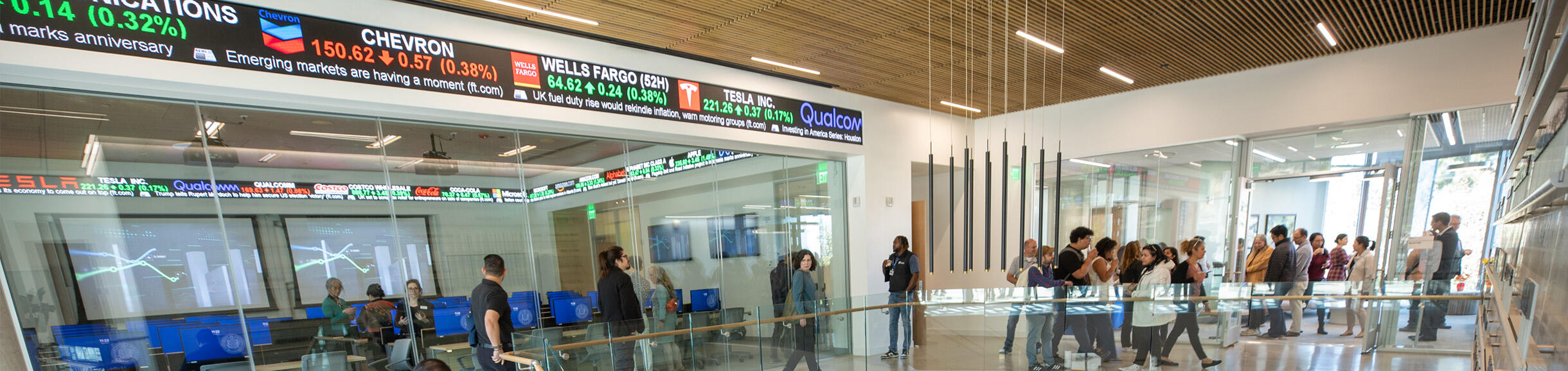 The modern atrium of the new UCR School of Business building with a large digital stock ticker and glass-walled classrooms.