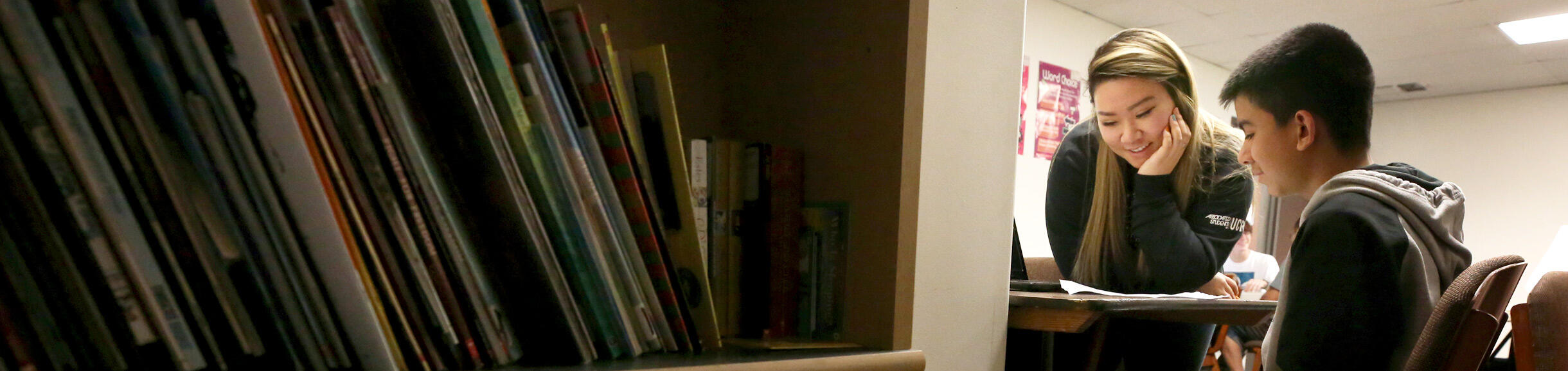 An educator sits at a desk helping a student with coursework. A full bookshelf is in the foreground of the classroom.