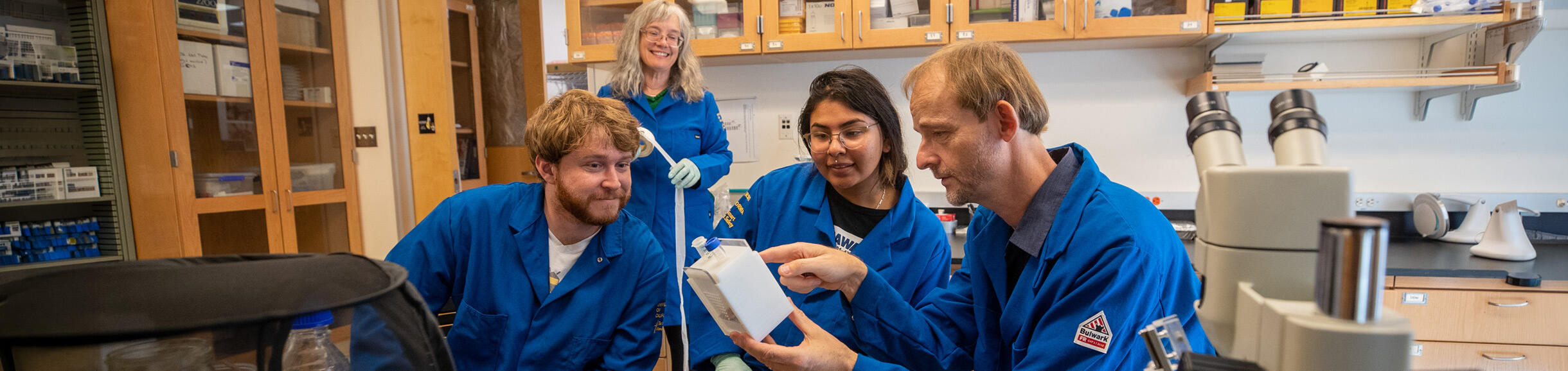 Four researchers in blue lab coats gather around a laboratory bench to examine a small, white scientific container. 