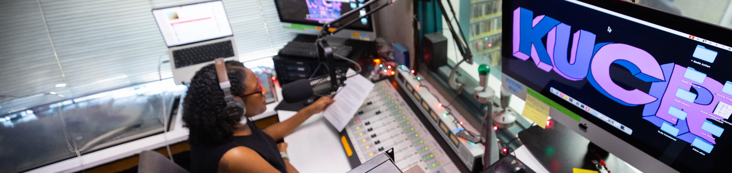 A woman with headphones speaking into a microphone in the "KUCR" radio studio, holding a script over a mixing console. 
