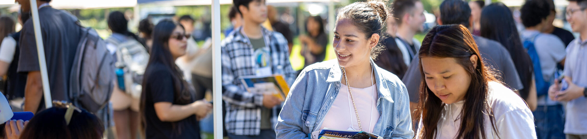Two female students look over a UC Riverside brochure at a sunny, outdoor recruitment fair.