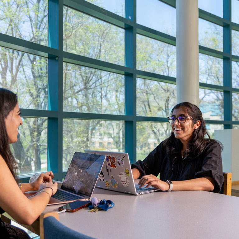 Two UCR students talk and work on laptops at a library table, with large windows showing the campus behind them.