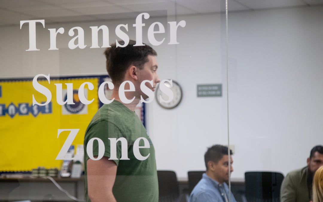 A group of students are seating in a conference room inside the Academic Resource Center (ARC). The view is looking through the window and there is a decal on the window that says "Transfer Success Zone."